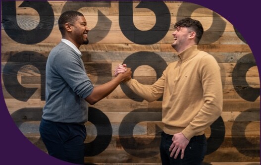 A young black man in a grey sweater shakes hands with a young white man in a tan sweater, in front of a wooden wall adorned with repeating Ally logos.