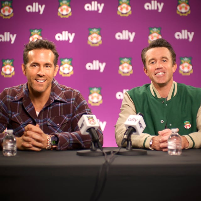 Ryan Reynolds and Rob McElhenney sit at a press conference table with Ally and Wrexham AFC logos on the wall behind them.