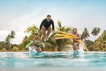 A man and two kids jumping into a pool with pool noodles