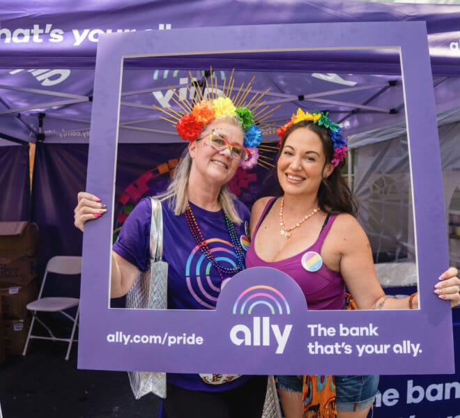 Two smiling women wearing rainbow floral headbands pose in a purple Ally Pride frame.