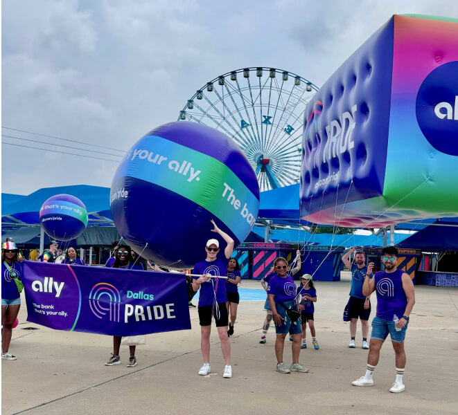 Excited parade participants show off their Ally Dallas Pride banner and balloons in front of a ferris wheel.