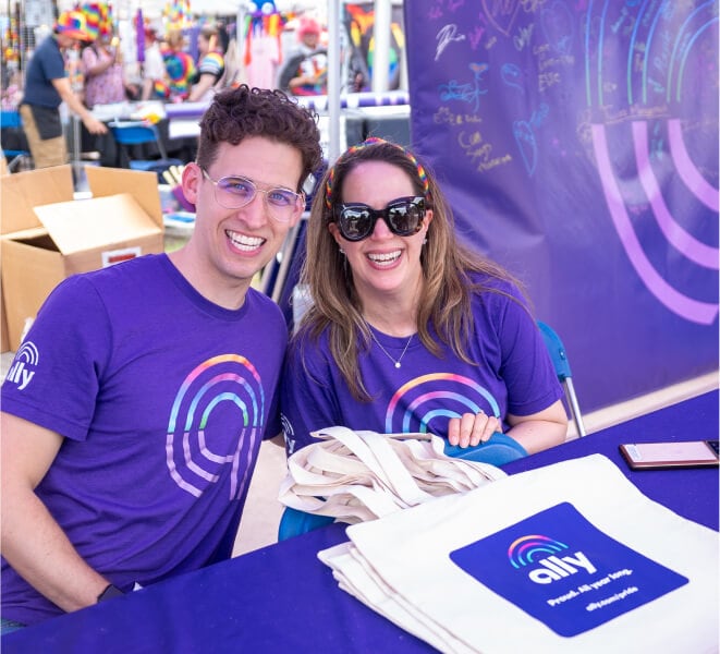 Two Ally Pride volunteers smile together behind the Ally booth.