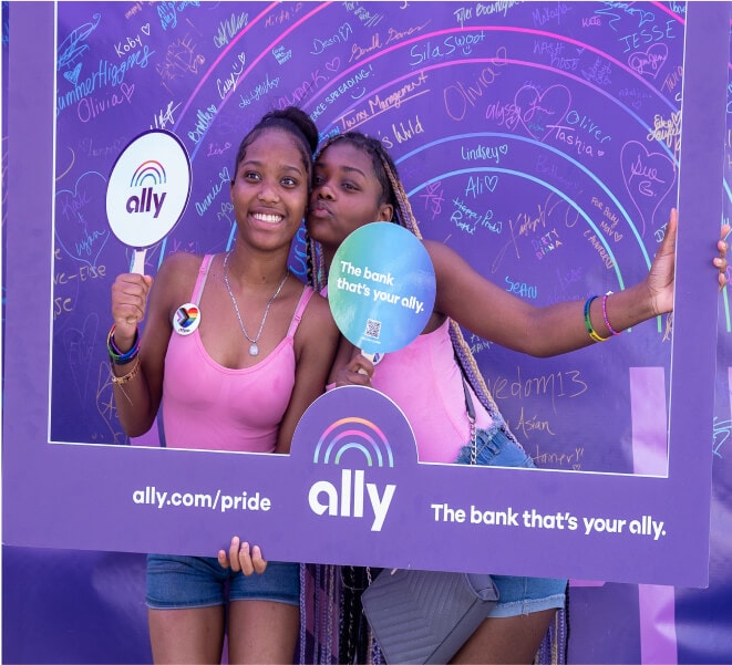 Two happy young women wearing matching pink tops pose with the Ally Pride frame and fans in front of the Allies Arc project.