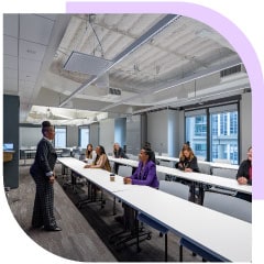 In a modern-looking boardroom, a black woman in a suit gives a presentation to people sitting at long white tables and taking notes.
