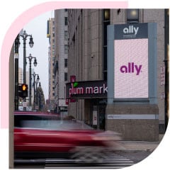On a busy city street, an electronic billboard displays the Ally logo on a lighter purple background as a red car zooms by.