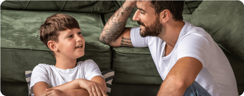 A child and a bearded man talk while sitting on the living room floor.