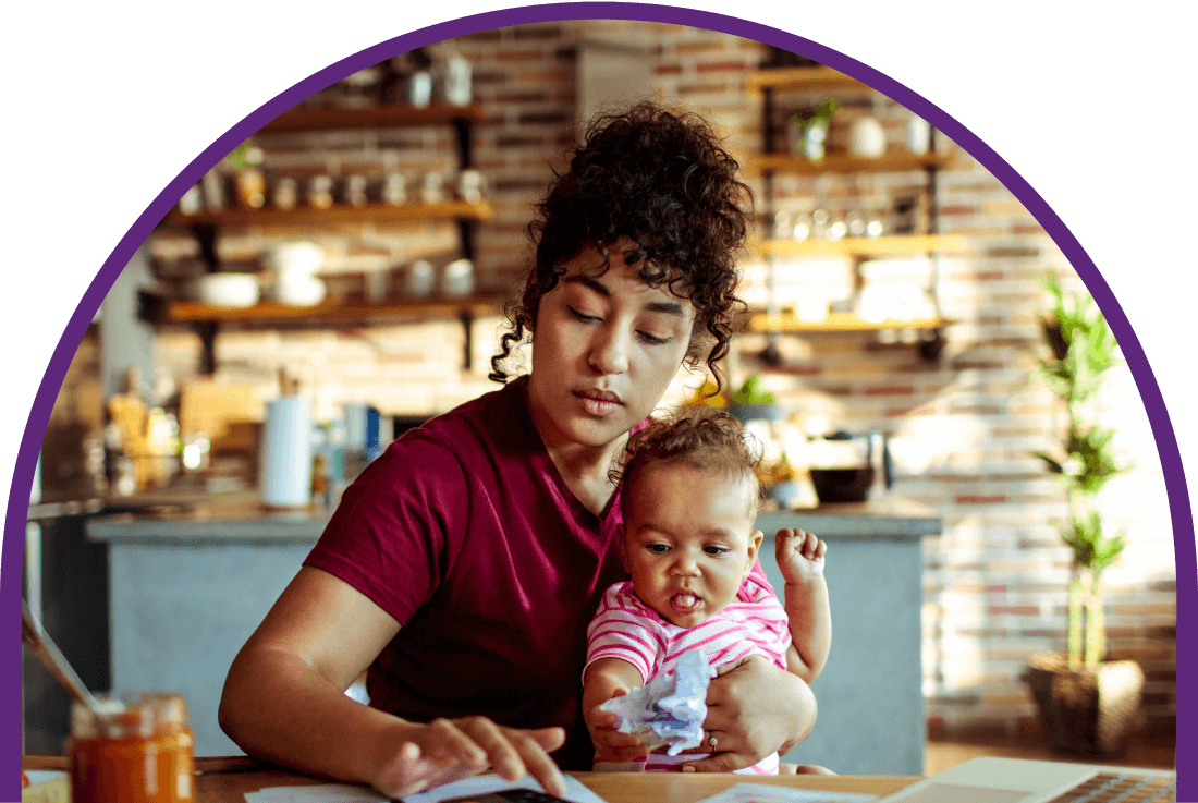 Curly-haired mother holds her child while doing bills at the kitchen table.