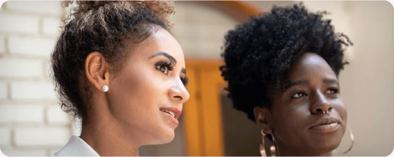 Two women with their curly hair done up are intrigued by something they see.