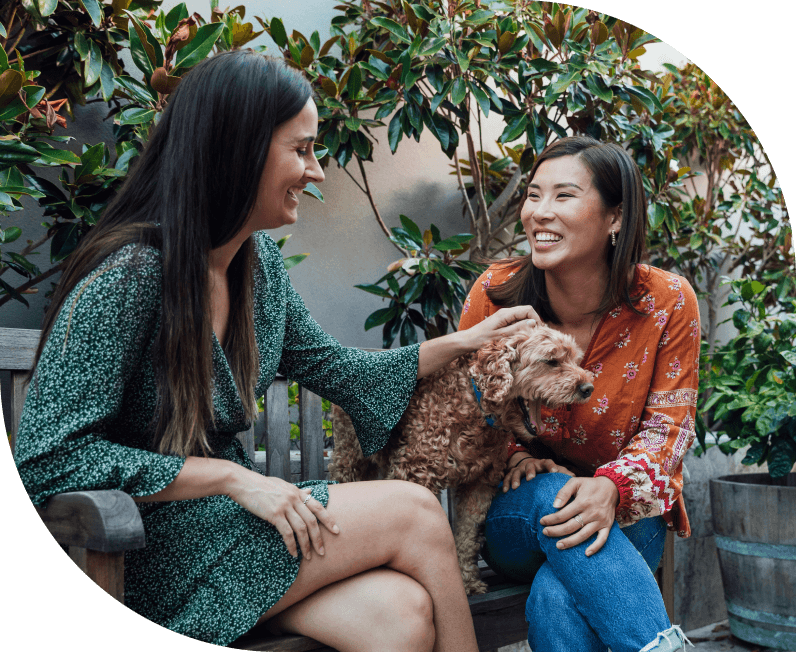 Two smiling women sit outside with a small dog