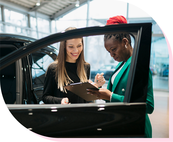 Two women standing behind an open car door in a dealership looking at a clipboard.