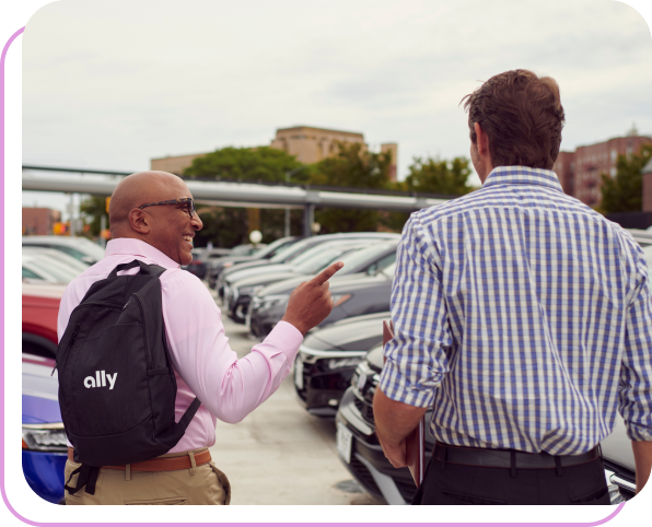 Two men, one with an ally backpack on, walking through a dealership lot full of cars.