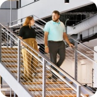 A white woman with red hair in a black shirt and tan pants and a white man with brown hair, a beard, a turquoise shirt and grey pants walk down a modern-looking flight of stairs at the Ally corporate office.