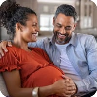 A pregnant brown-skinned woman in a dark orange blouse smiles as a smiling dark skinned man in a grey shirt rests a hand on her stomach.