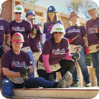 A group of people in purple Ally volunteer t-shirts and hard hats pose in front of the wooden frame of a house under construction