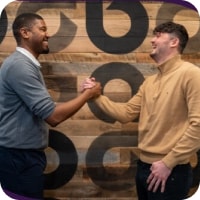 A young black man in a grey sweater and black pants shakes hands with a young white man in a tan sweater and black pants in front of a wooden wall with repeating lowercase a Ally logos on it.