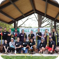 A large group of ally employees are outdoors, wearing Ally volunteer shirts and holding shovels, standing in front of a sign that says, ‘Tree Planting’.