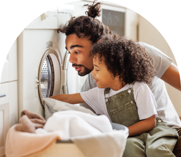 An Afro-Latino man, with messy-bun-styled hair, and an Afro-Latino boy, dressed in overalls, load laundry into the washer. 