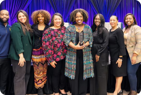 Eight women, all of different ethnic backgrounds, stand in front of a purple curtain.