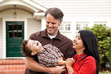 A father, mother and child laugh in front of their new home on a sunny day.