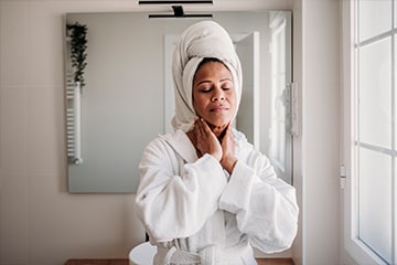 A woman rubbing her neck while wearing a robe with her hair wrapped in a towel.