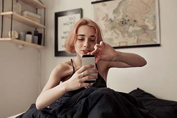 Woman sits on her bed in her room, looking at her phone.