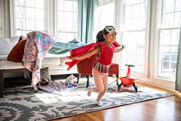 Little girl wearing a cape and a mask running through living room playing.