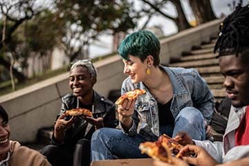 Friends sitting on a staircase in a park and eating pizza.